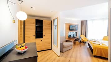 A modern hotel room with a wooden floor, a dining area with a yellow table and chairs, a bed with a yellow bedspread, a TV mounted on the wall, and a fruit bowl on the counter.
