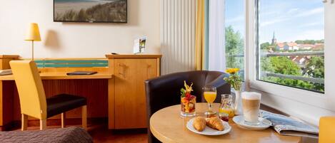 A hotel room with a dining area, a bed, a desk, a chair, a TV, and a window with a view of a town.
