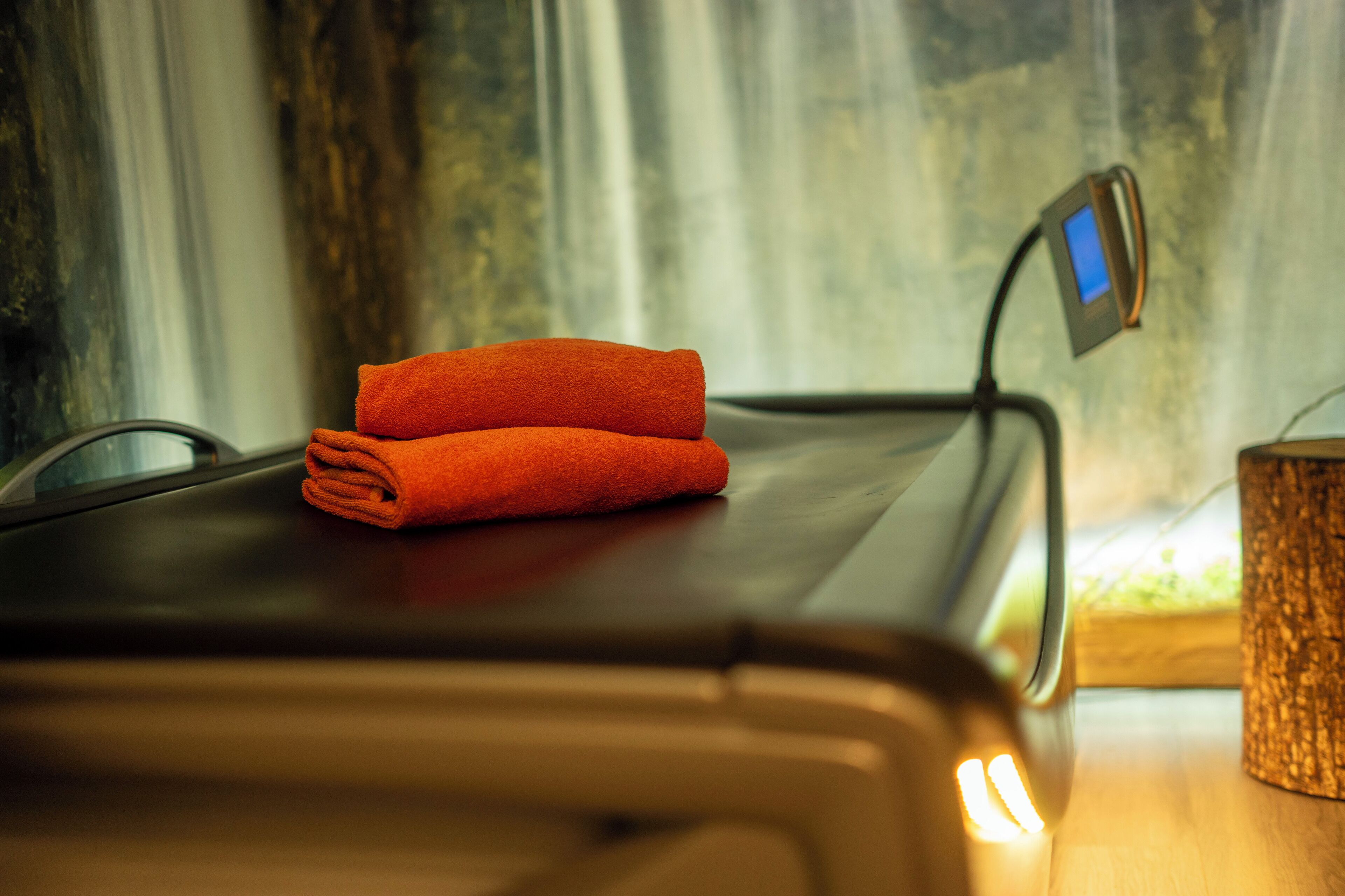 A massage table with a folded orange towel on it, a digital device on the table, and a wooden side table with a textured candle holder.