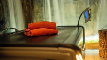Une table de massage sur laquelle est posée une serviette orange pliée, un appareil numérique sur la table, et une petite table en bois avec un porte-bougie texturé.