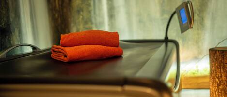 A massage table with a folded orange towel on it, a digital device on the table, and a wooden side table with a textured candle holder.