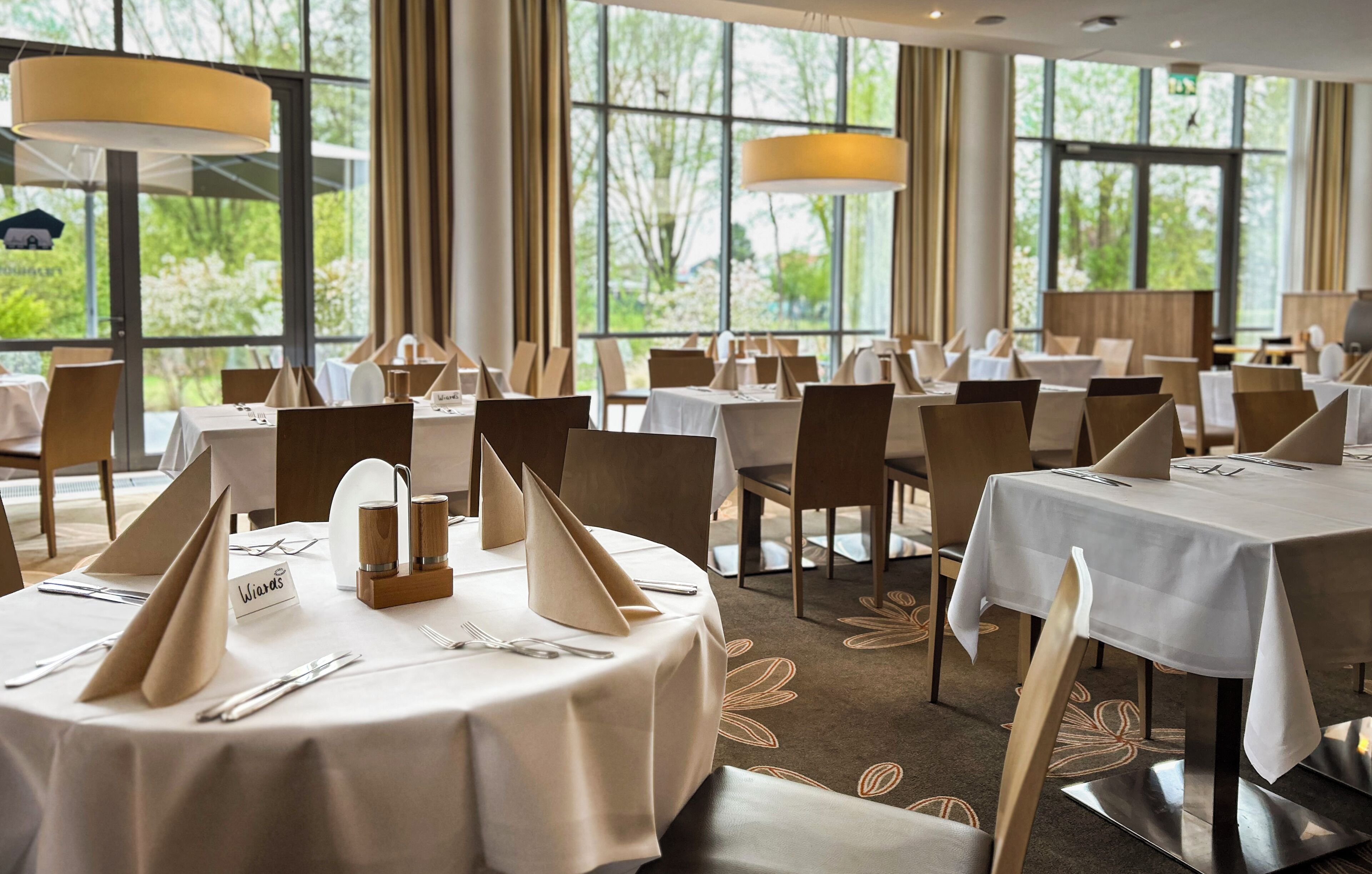 A restaurant with round tables set for dining, large windows, and a view of greenery outside.