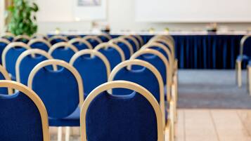 Filas de sillas azules con armazón de madera en una sala de conferencias con una larga mesa y una planta al fondo.
