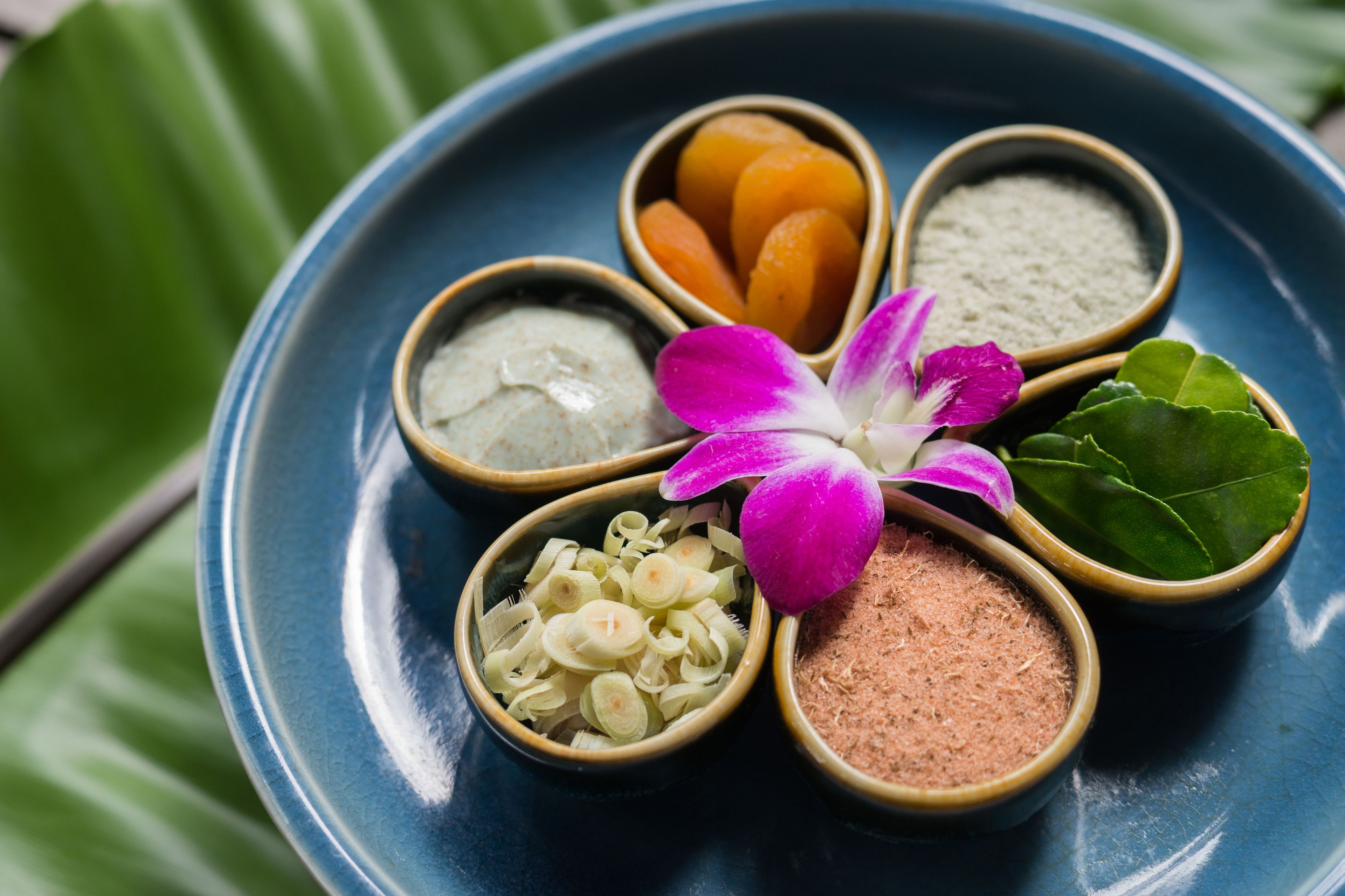 A blue plate with six small bowls containing different ingredients, a purple flower, and a green leaf.