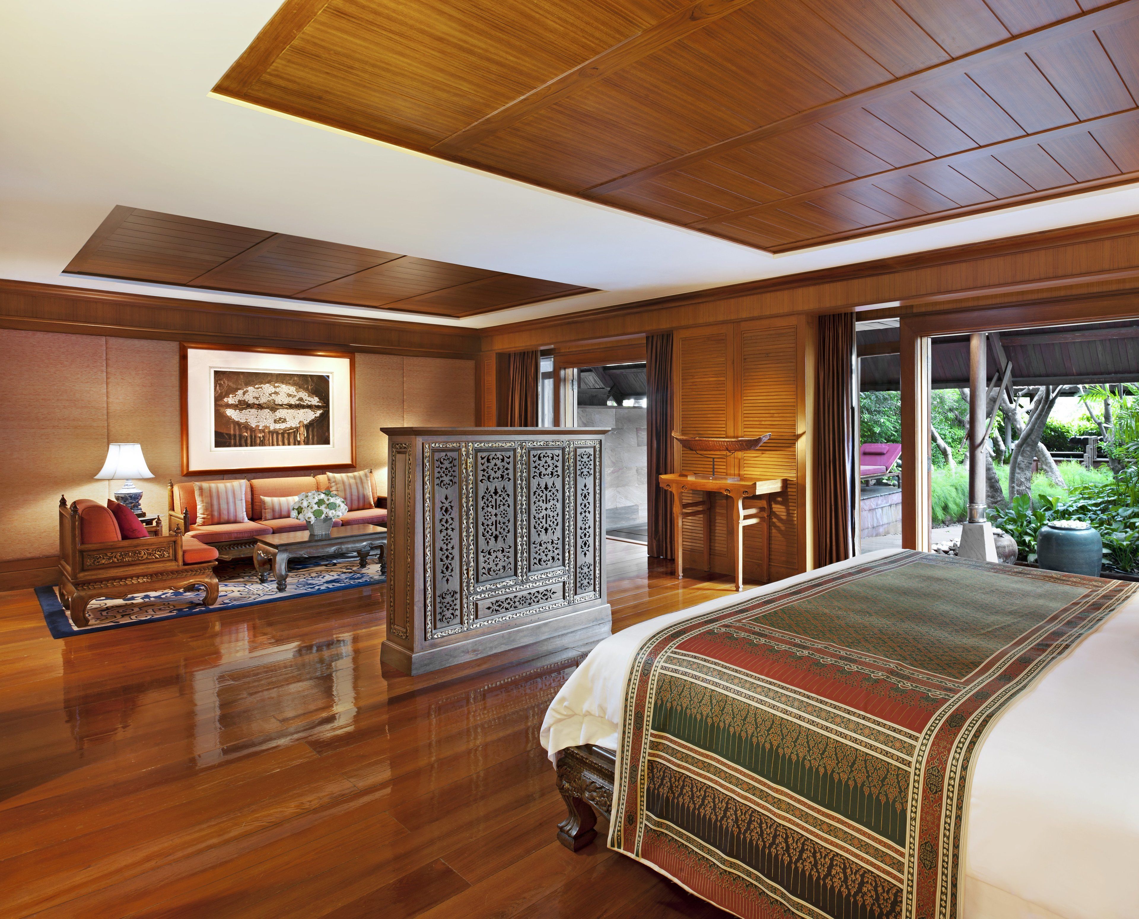 A spacious hotel room with a large bed, a sitting area with a sofa and armchairs, a wooden cabinet, and a view of the outdoors through large sliding doors.