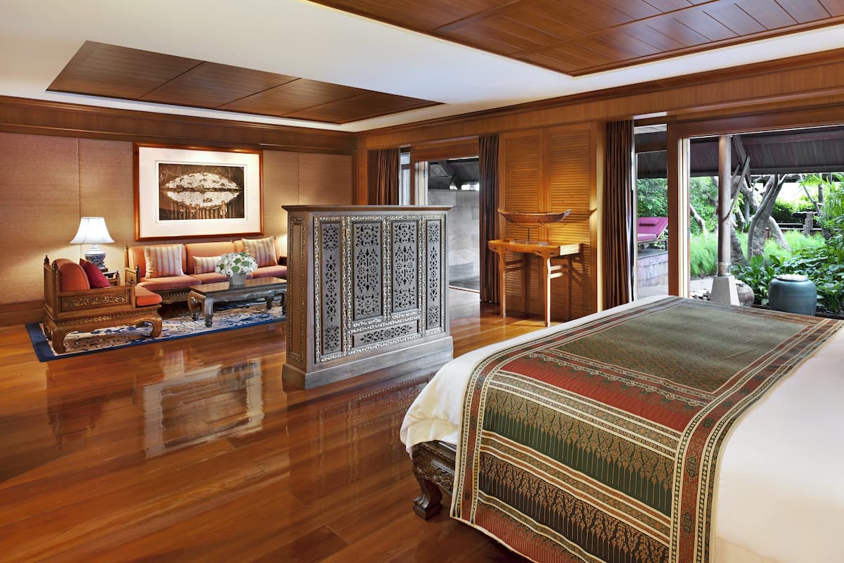 A spacious hotel room with a large bed, a sitting area with a sofa and armchairs, a wooden cabinet, and a view of the outdoors through large sliding doors.