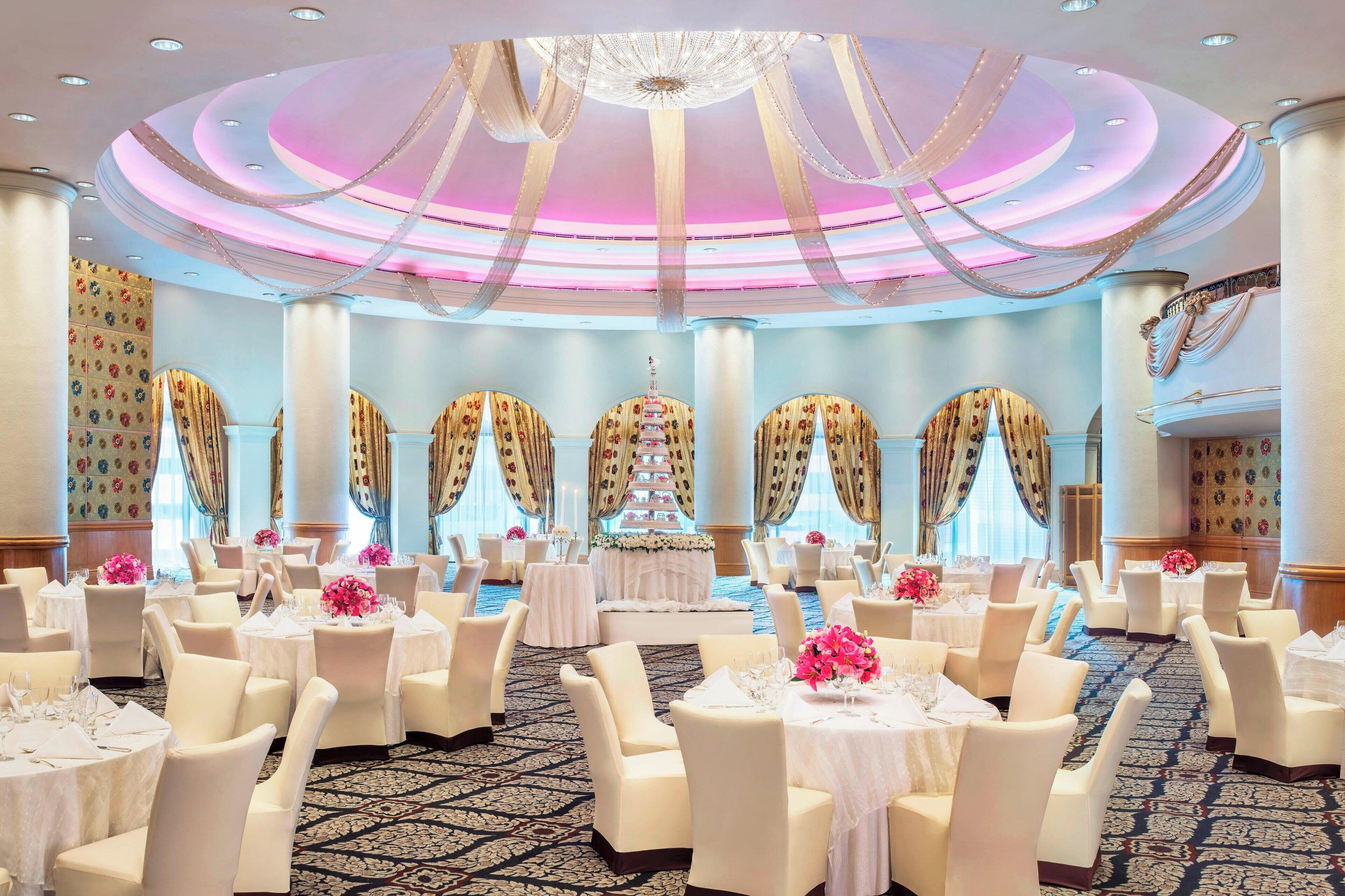 A banquet hall with round tables, chairs, and a tiered cake display.