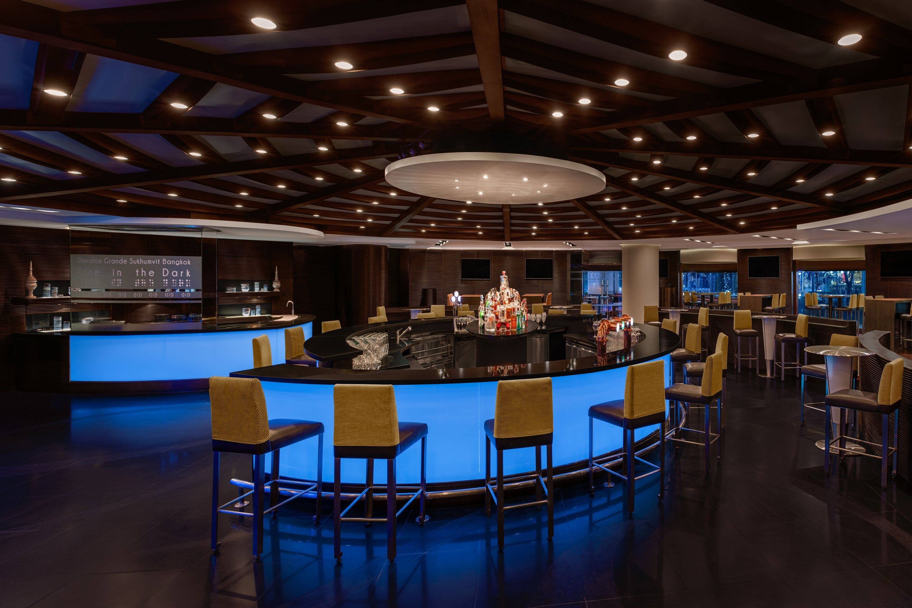 En moderne bar med en central bardisk, flere siddepladser og et loft med indbygget belysning.
