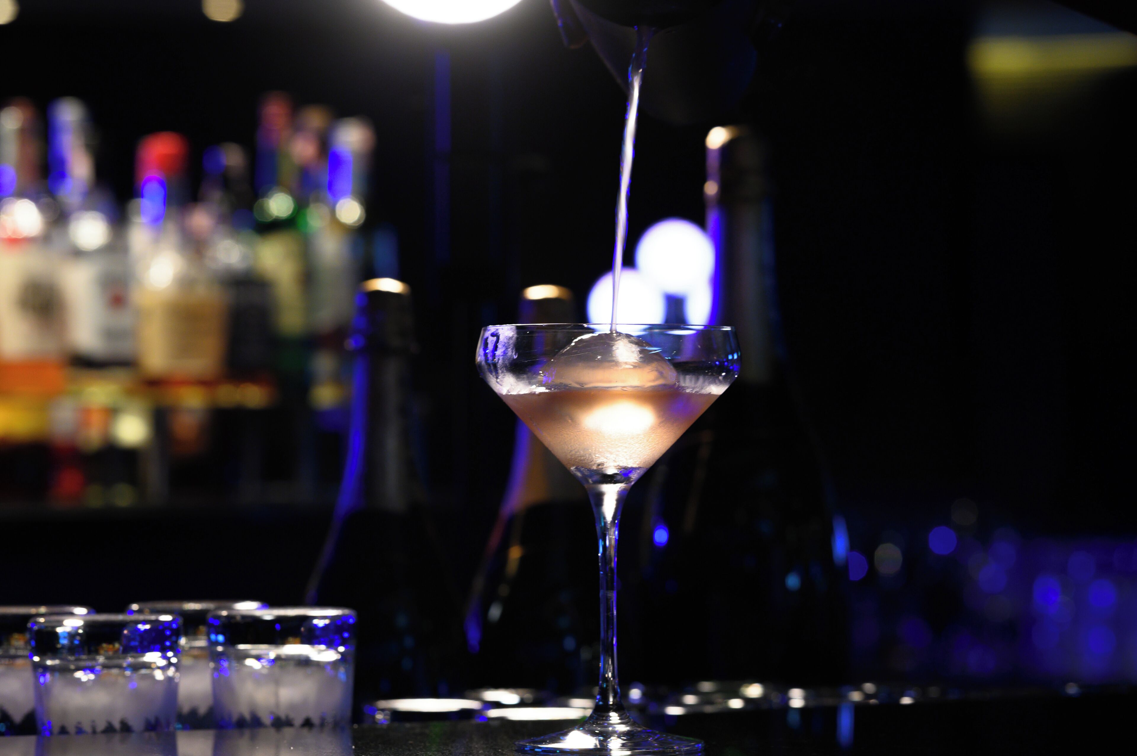 Un verre à martini contenant un liquide transparent qui y est versé, posé sur le comptoir d’un bar, avec des bouteilles en arrière-plan.