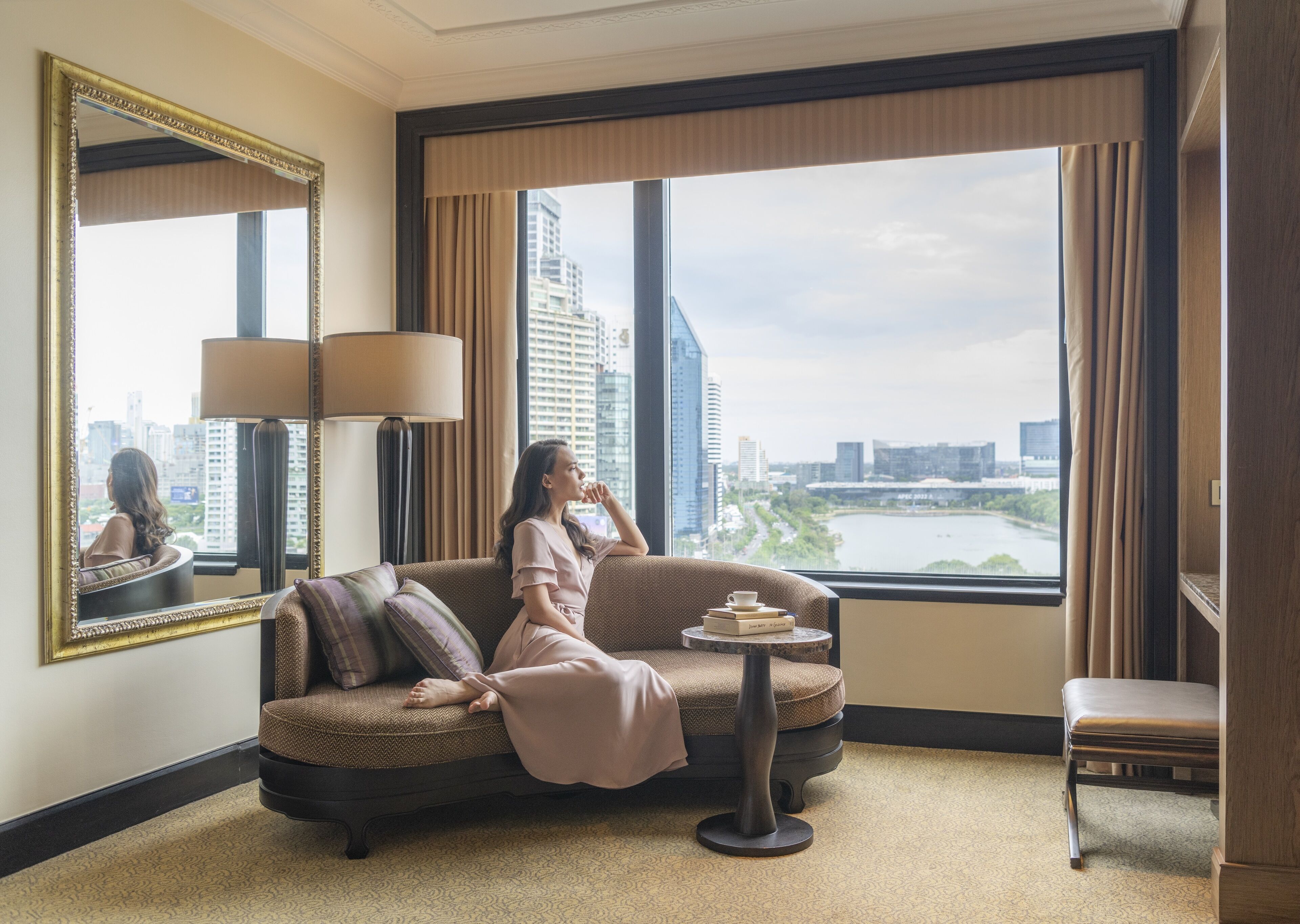 A hotel room with a large window, a sofa, a small table with books, and a chair.