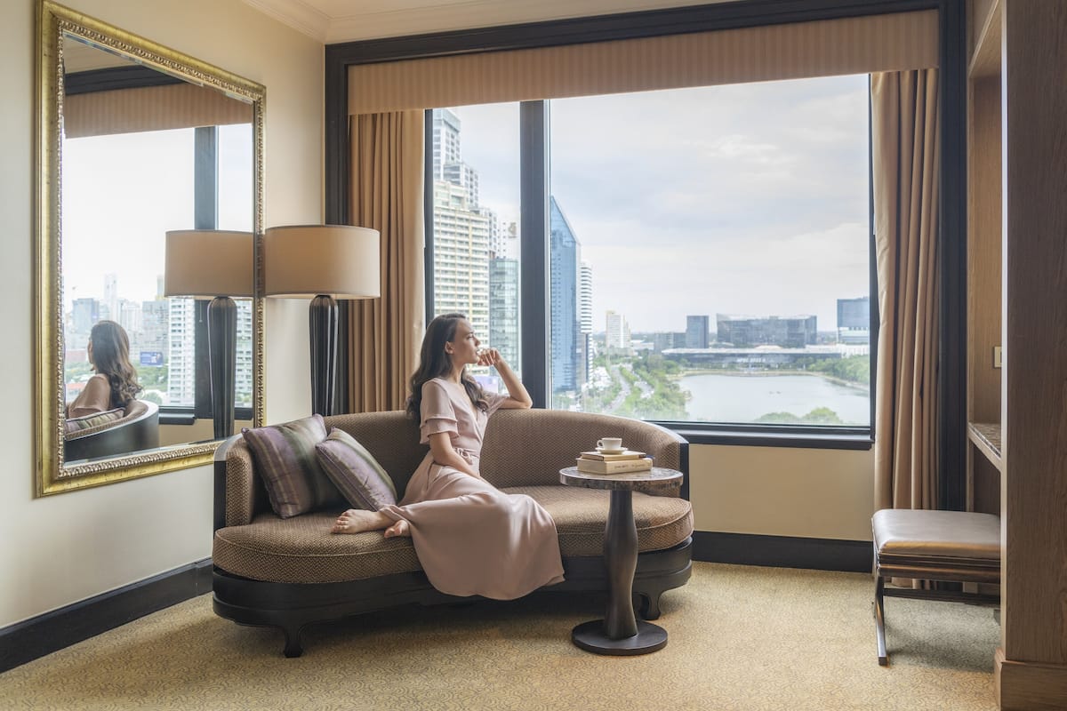 A hotel room with a large window, a sofa, a small table with books, and a chair.