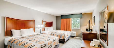 A hotel room with two beds, a desk with a lamp, a mirror, and a window with curtains.