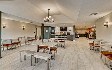 A spacious dining area with multiple tables and chairs, a central chandelier, and a kitchen in the background.