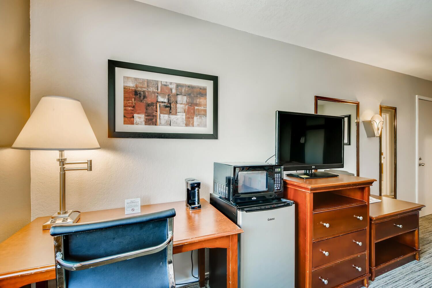 A hotel room with a desk, chair, coffee maker, microwave, television, and a framed picture on the wall.