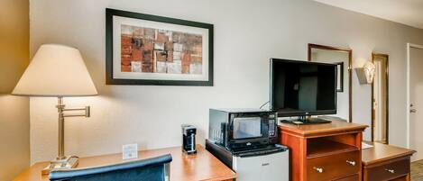 A hotel room with a desk, chair, coffee maker, microwave, television, and a framed picture on the wall.