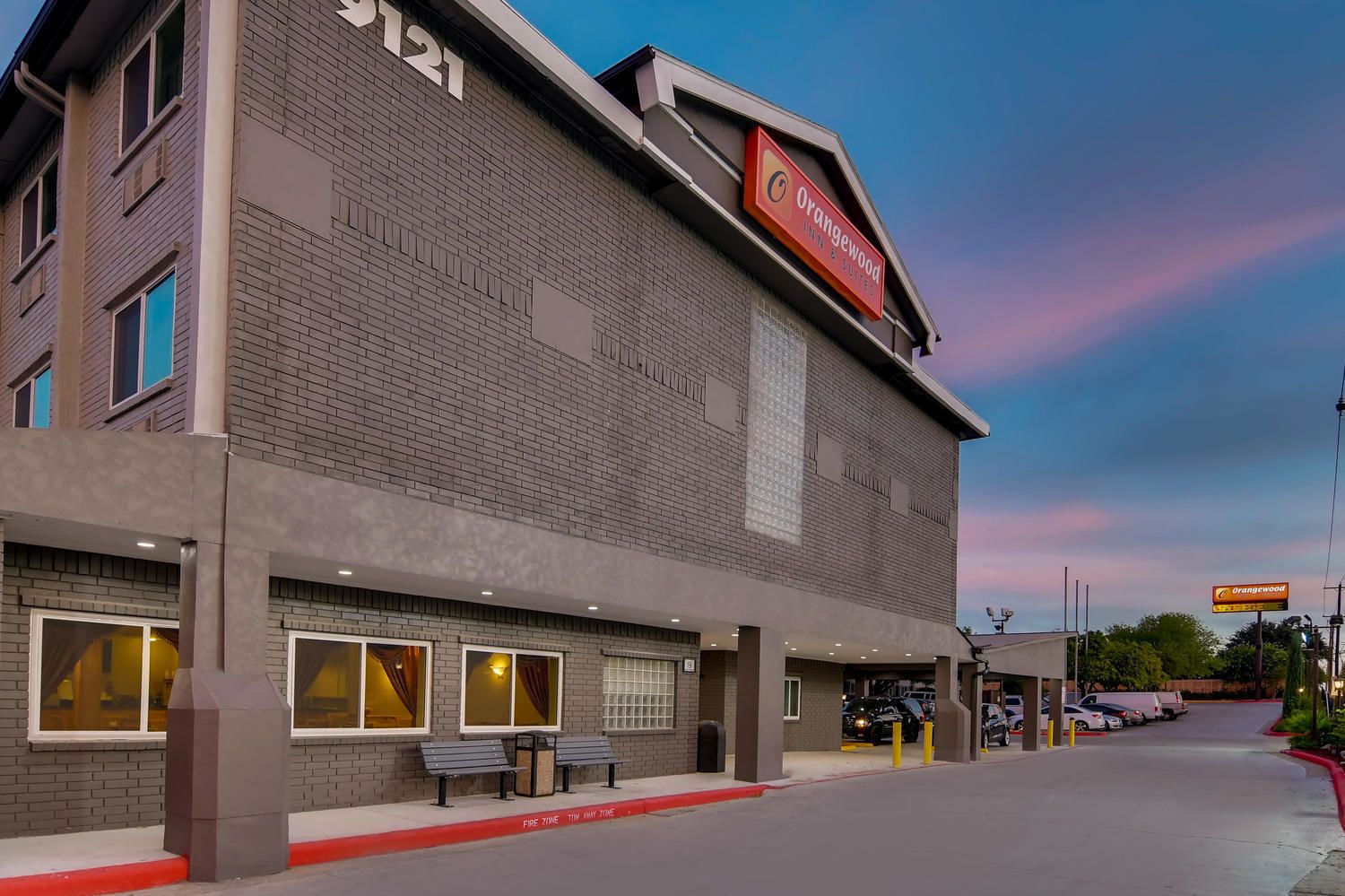 A hotel with a brick facade, a prominent Orangewood Inn & Suites sign, and a fire lane marked on the sidewalk.