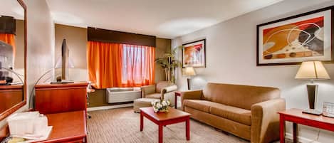 A hotel room with a sofa, a coffee table, a desk, a lamp, and a large window with orange curtains.