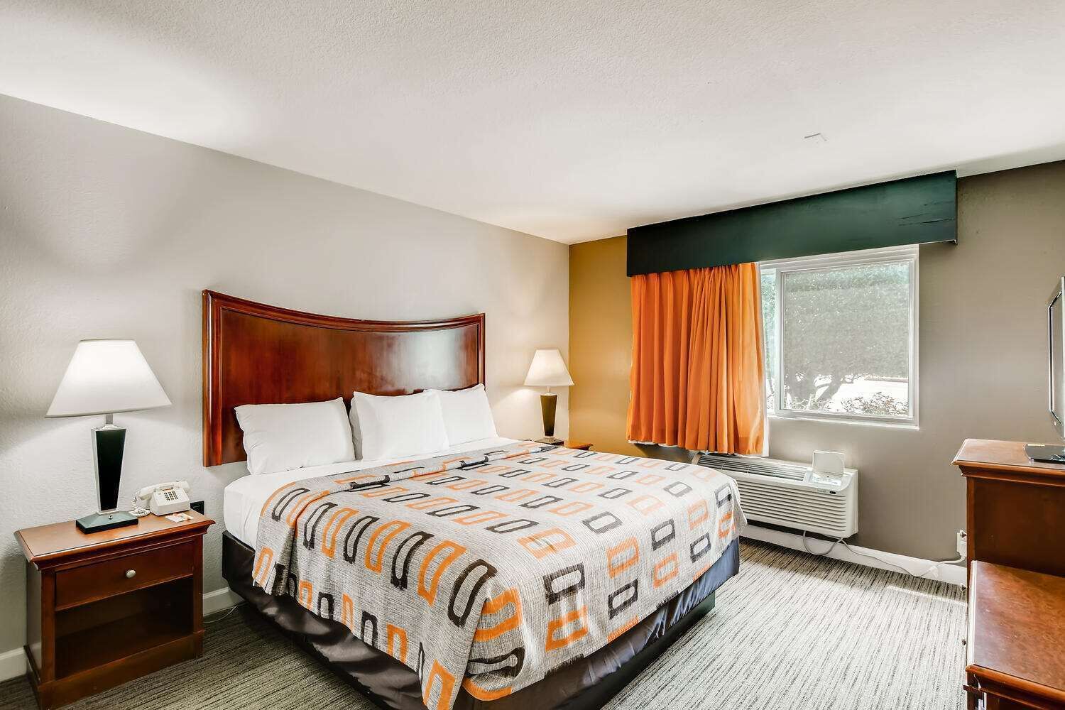 A hotel room with a bed, two bedside tables, a desk, a chair, a window with curtains, and a wall-mounted TV.