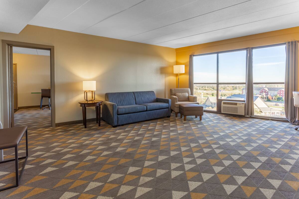A hotel room with a blue sofa, a chair, a small table, and a large window offering a view of the outside.