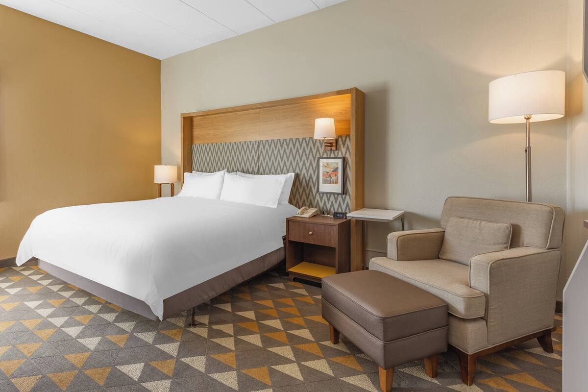 A hotel room with a large bed, a nightstand with a lamp, a chair, and a small table.