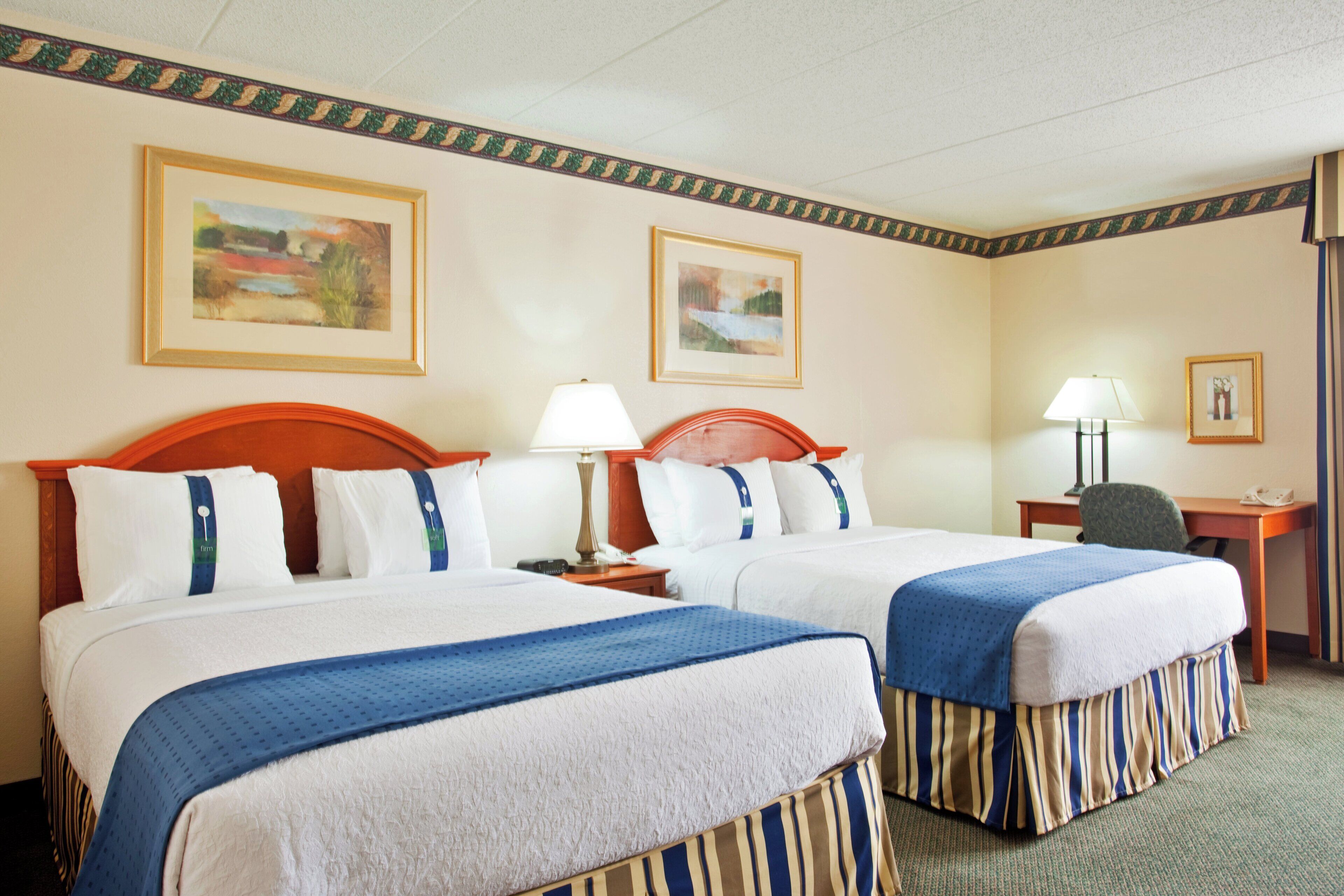 A hotel room with two beds, a desk, a chair, and two paintings on the walls.