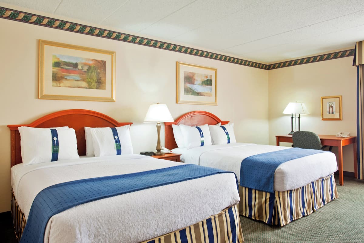 A hotel room with two beds, a desk, a chair, and two paintings on the walls.