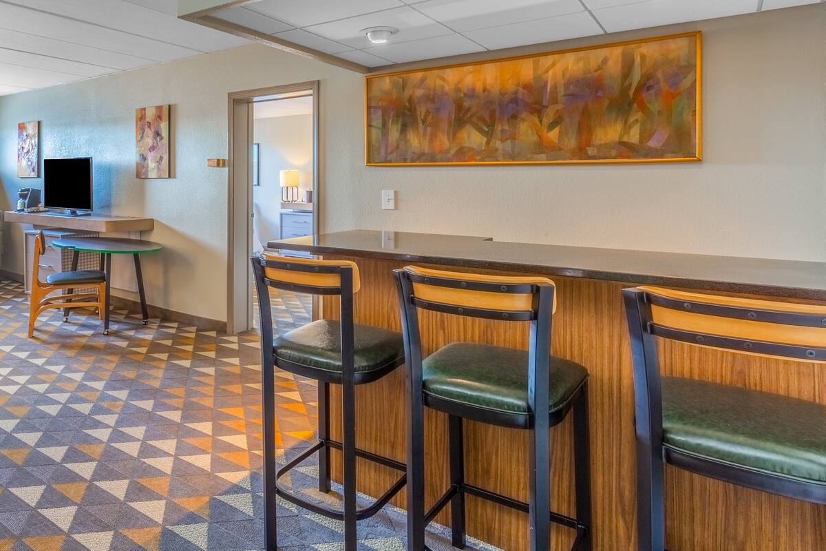 A bar area with wooden bar stools and a counter. There are two paintings on the wall and a television on a stand.