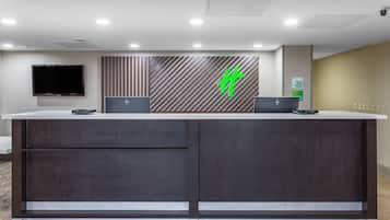 Un mostrador de recepción con diseño moderno, que incluye un televisor de pantalla plana grande, dos computadoras portátiles y un logotipo verde en la pared.