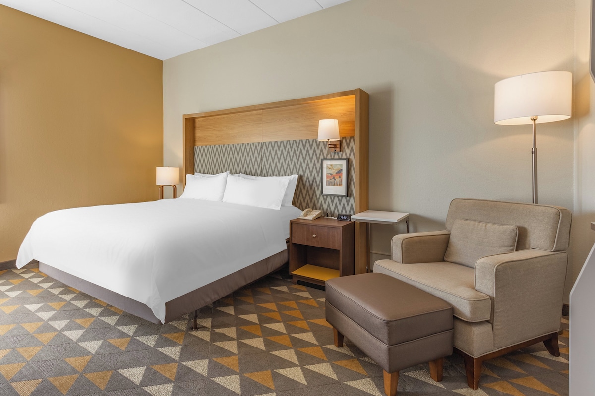 Una habitación de hotel con una cama grande, una mesita de noche con lámpara, una silla y una pequeña mesa.