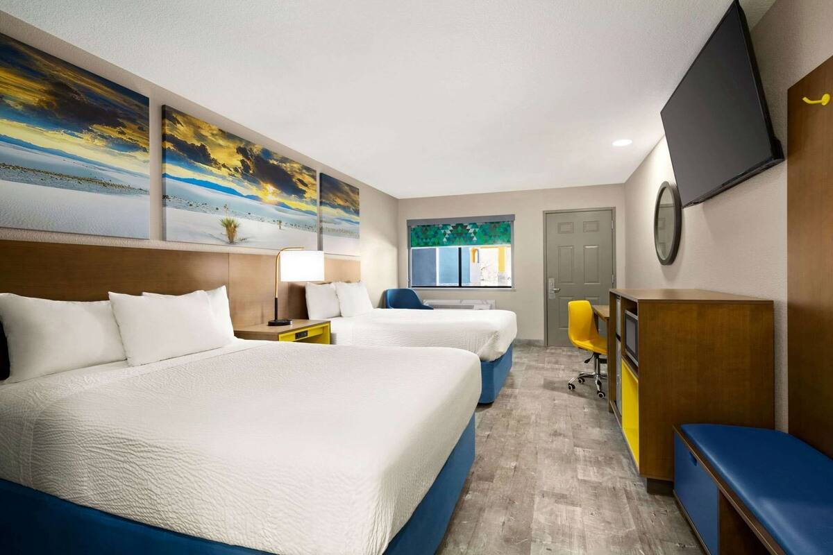 A hotel room with two beds, a desk, a chair, a TV, and artwork on the walls.
