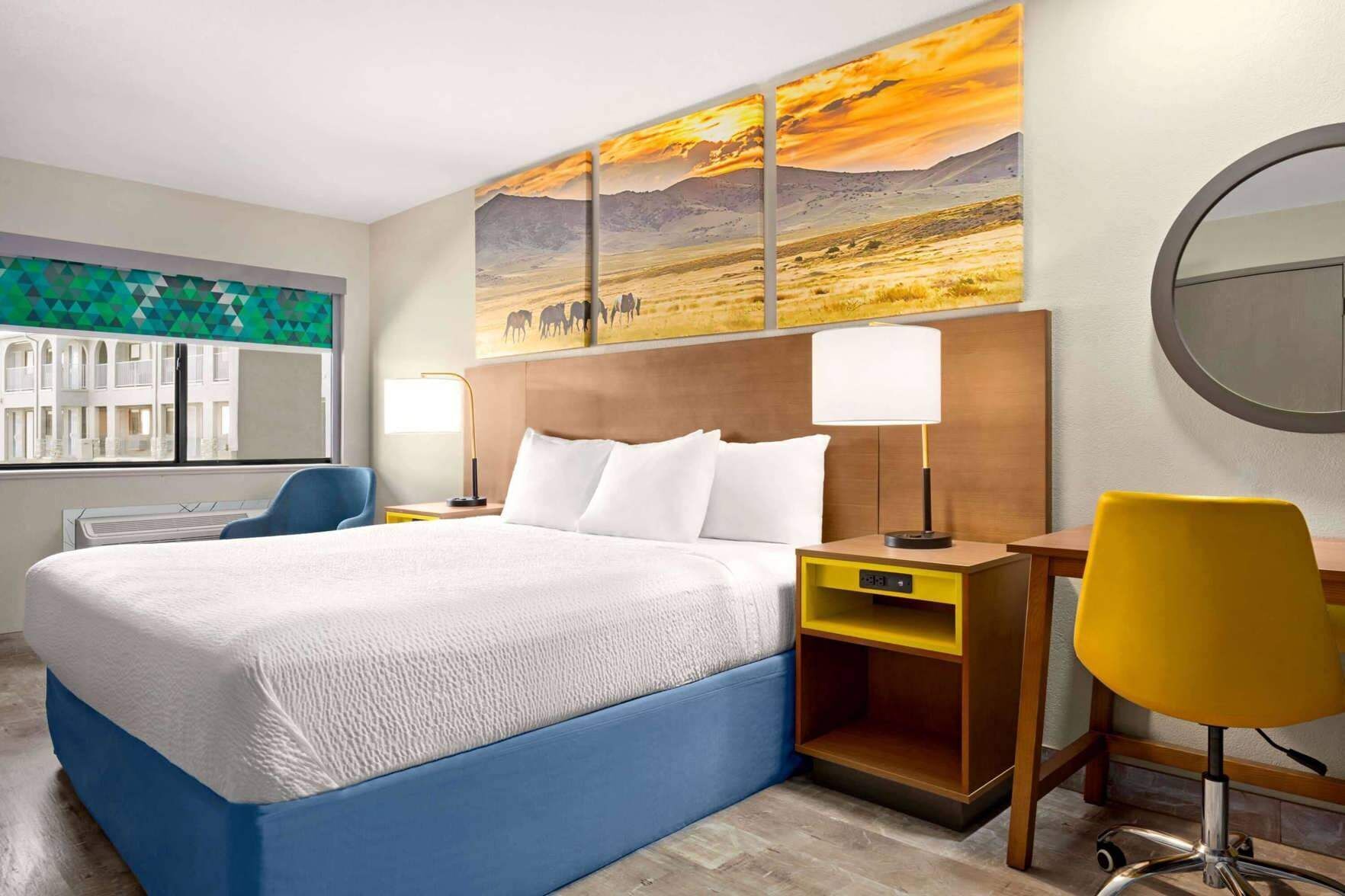 A hotel room with a bed, a desk, a chair, a lamp, and a large painting on the wall.