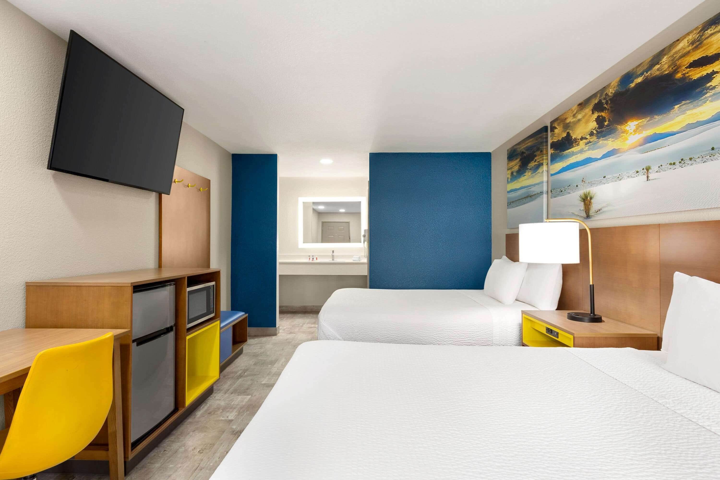 A hotel room with a bed, a desk with a TV, a small kitchenette, and a bathroom visible through an open door.