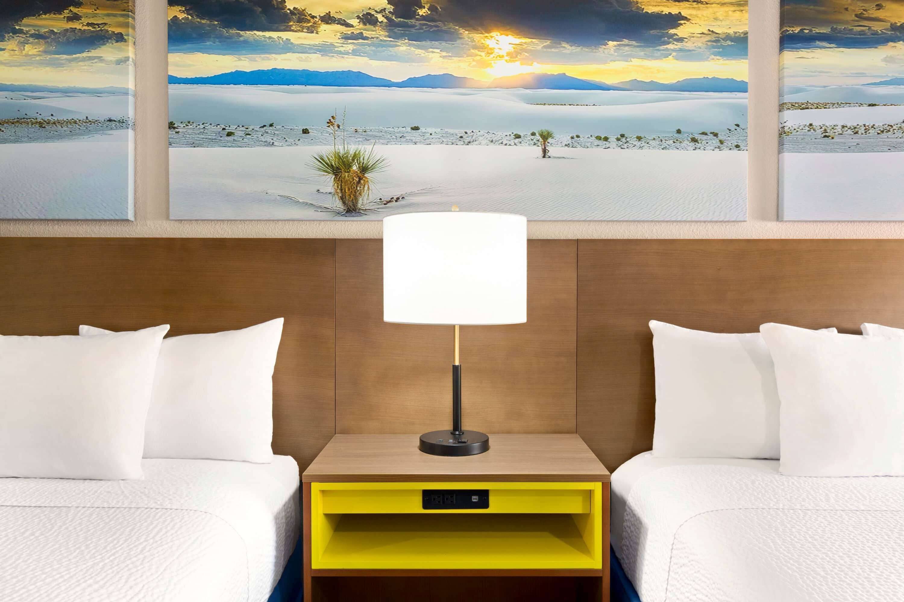 A hotel room with two beds, a nightstand with a lamp, a yellow storage drawer, and a large window with a desert landscape view.