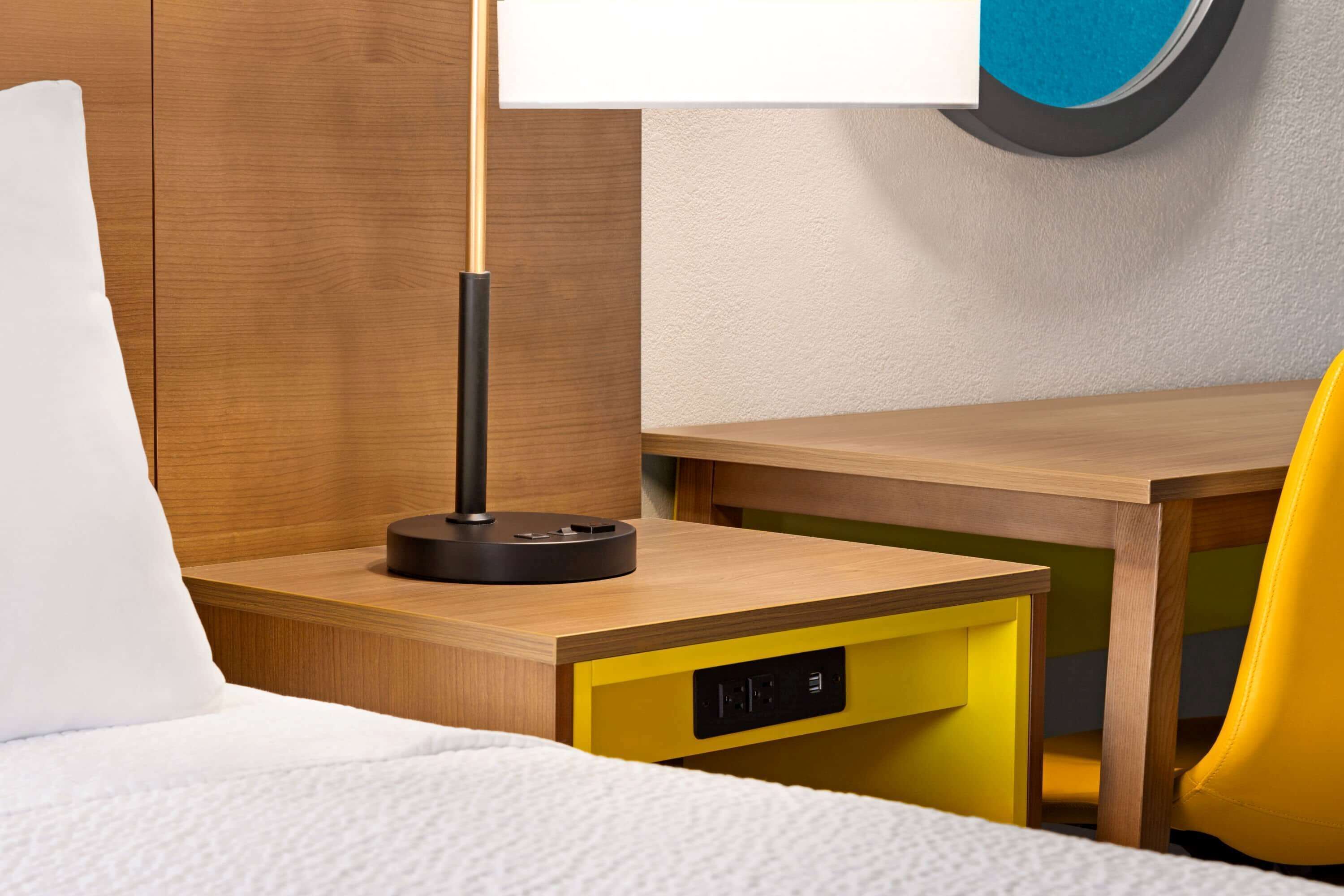 Una habitación de hotel con cama, escritorio con lámpara, una silla amarilla y un reloj de pared.