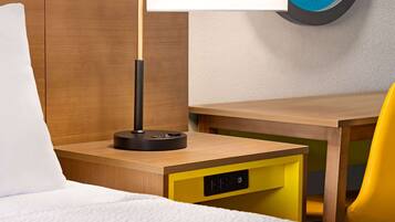 Una habitación de hotel con cama, escritorio con lámpara, una silla amarilla y un reloj de pared.