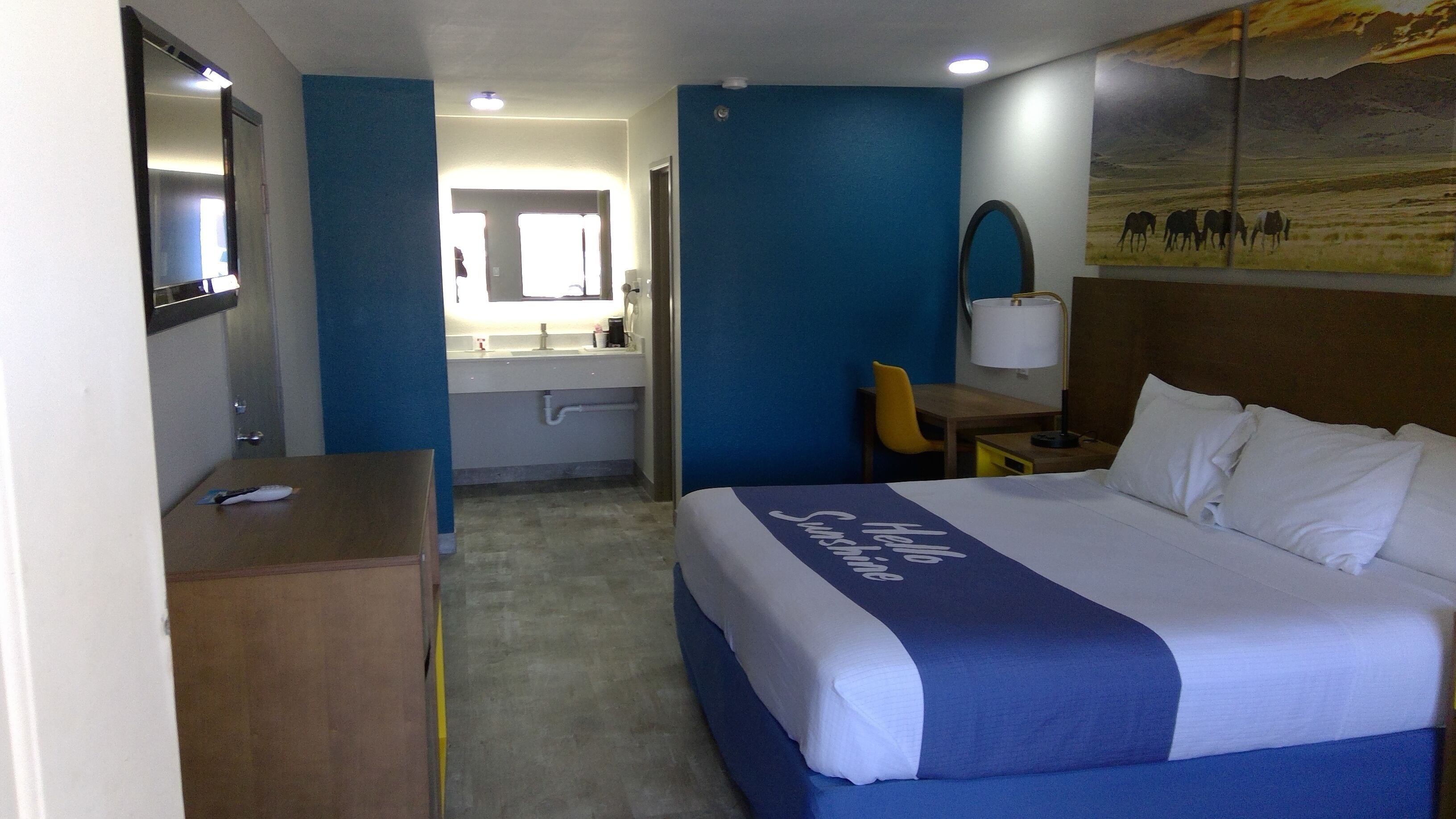 A hotel room with a bed, a desk, a chair, a mirror, a bathroom with a sink and toilet, and a painting on the wall.