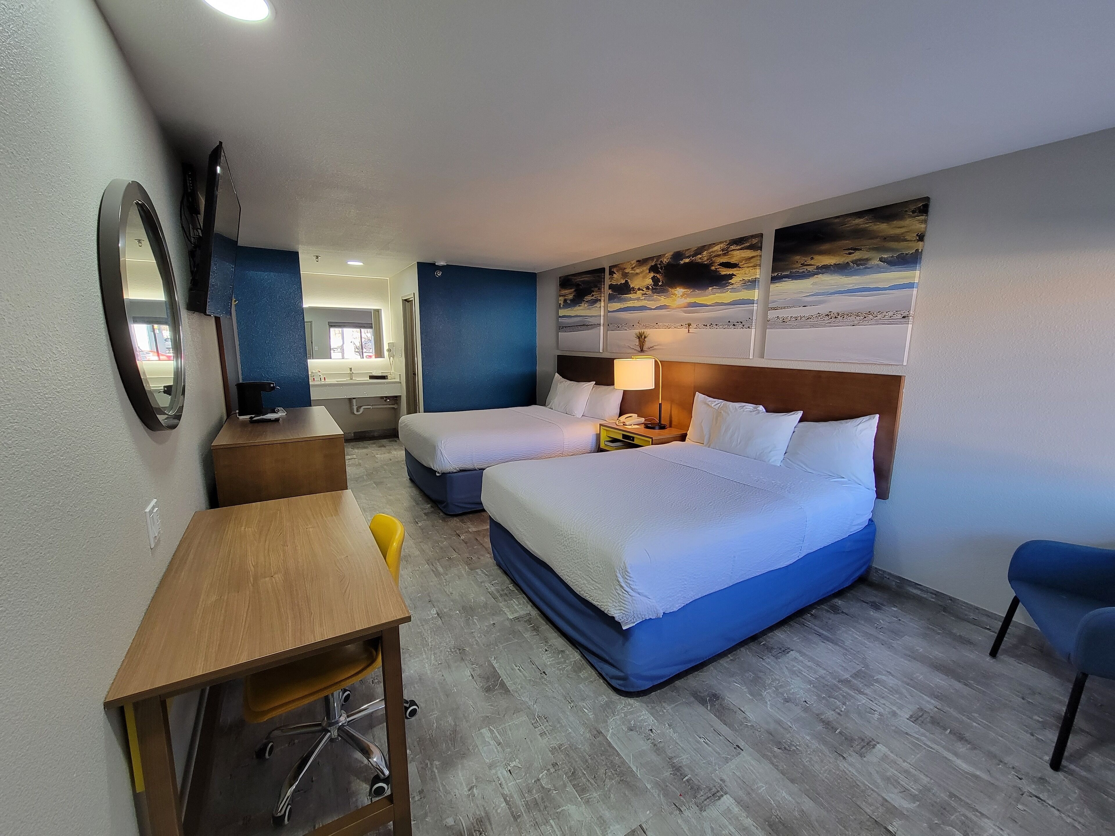 Une chambre d’hôtel avec deux lits, un bureau, une chaise, un miroir, une table de chevet avec une lampe et des œuvres d’art accrochées au mur.