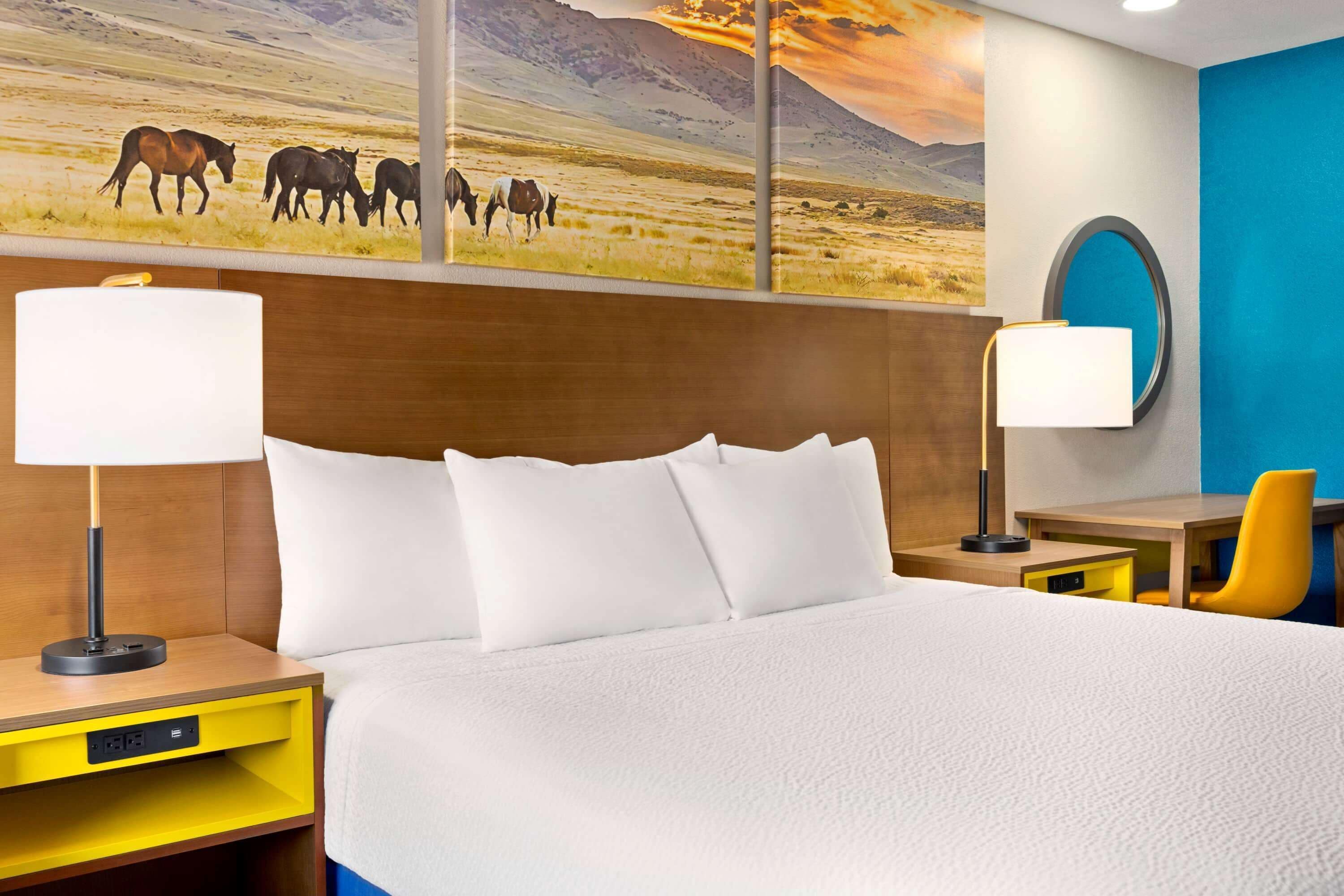 A hotel room with a large bed, two bedside tables, a desk, a chair, and a painting of horses on the wall.