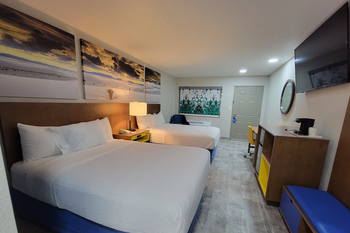 A hotel room with two beds, a desk, a chair, a coffee machine, and artwork on the walls.