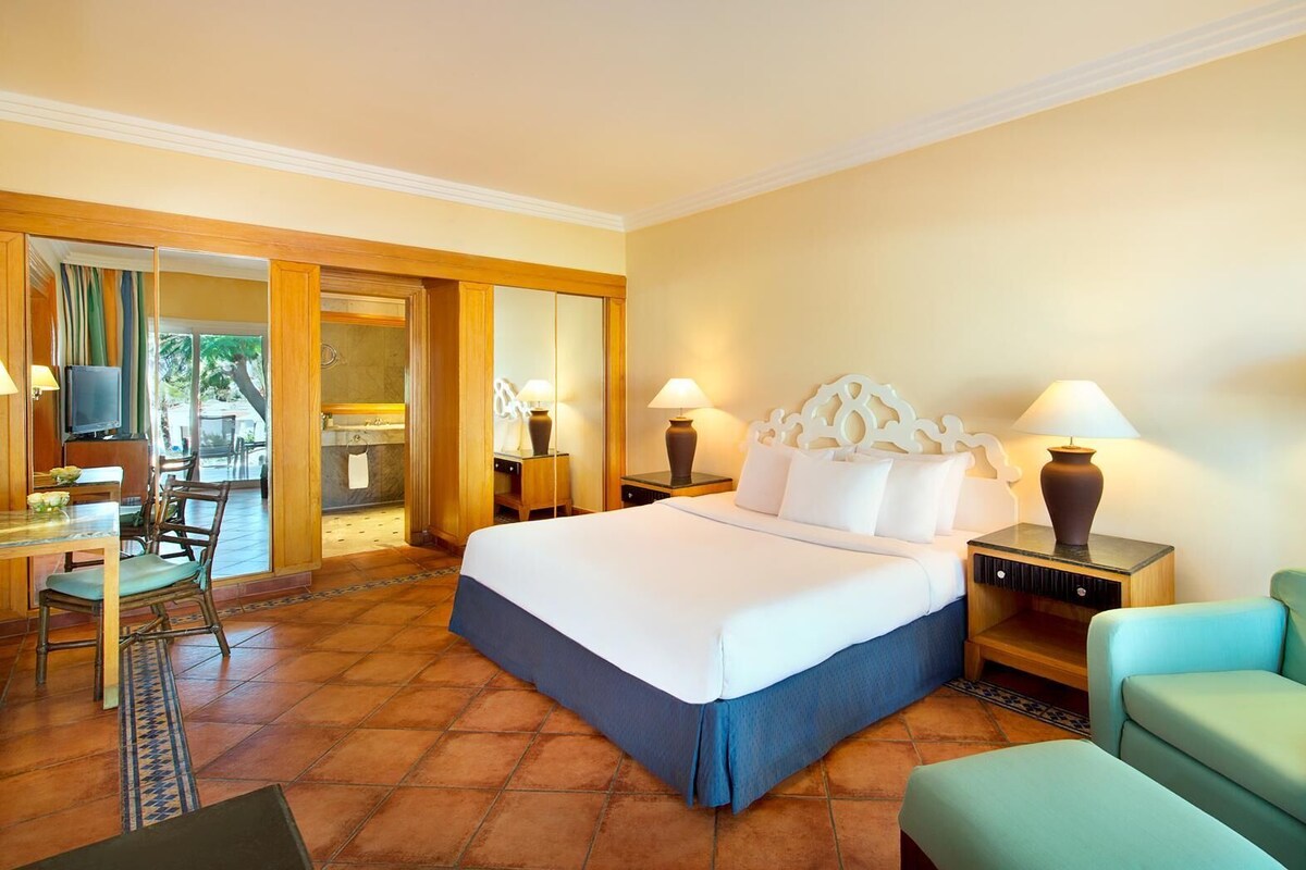 Habitación de hotel con cama, escritorio, silla, televisión y baño visible a través de una puerta de cristal.