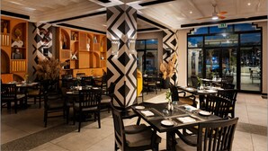 Um restaurante com interior moderno, apresentando padrões geométricos em preto e branco nas paredes e no teto, móveis de madeira e vista para a rua.