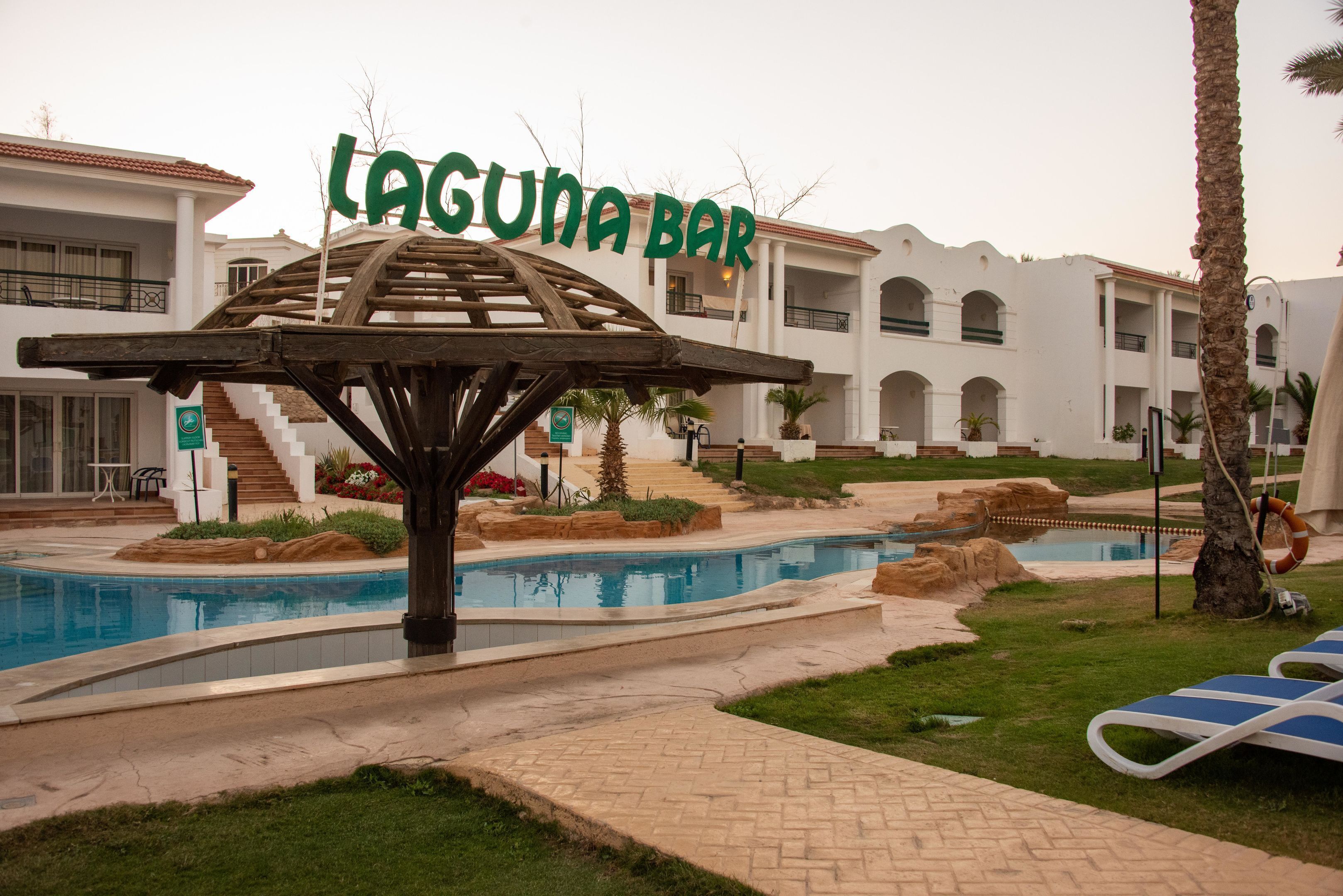 A pool area with a wooden structure labeled 'Laguna Bar,' a swimming pool, and a building with multiple balconies in the background.