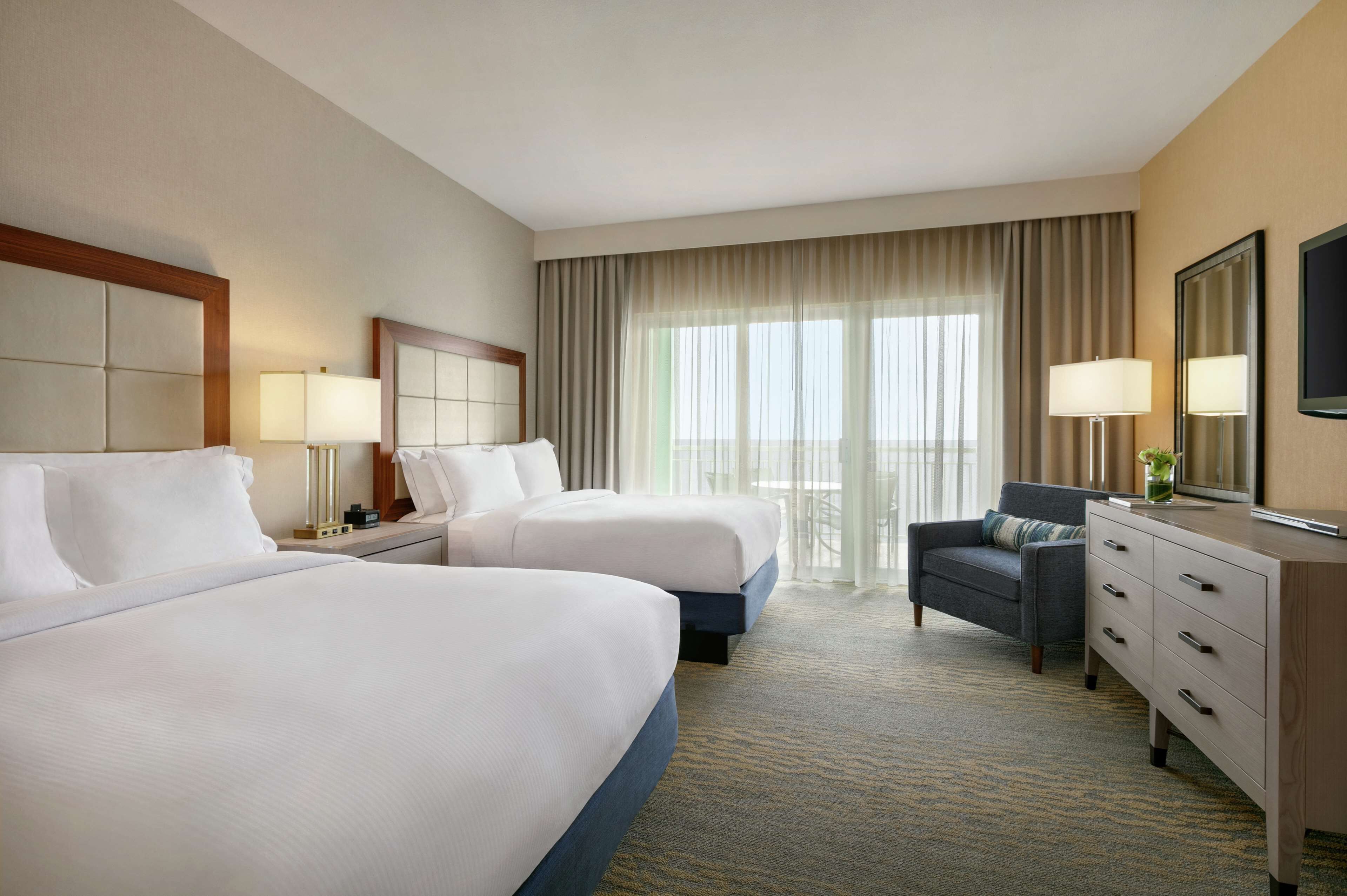A hotel room with two beds, a desk, a chair, and a large window with curtains.