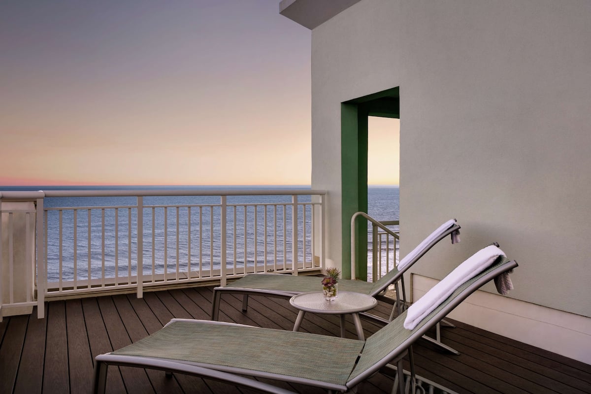 Un balcon avec deux fauteuils, une petite table et une vue sur l’océan au coucher du soleil.