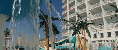 A hotel pool area with a water fountain, palm trees, and a multi-story building with balconies.