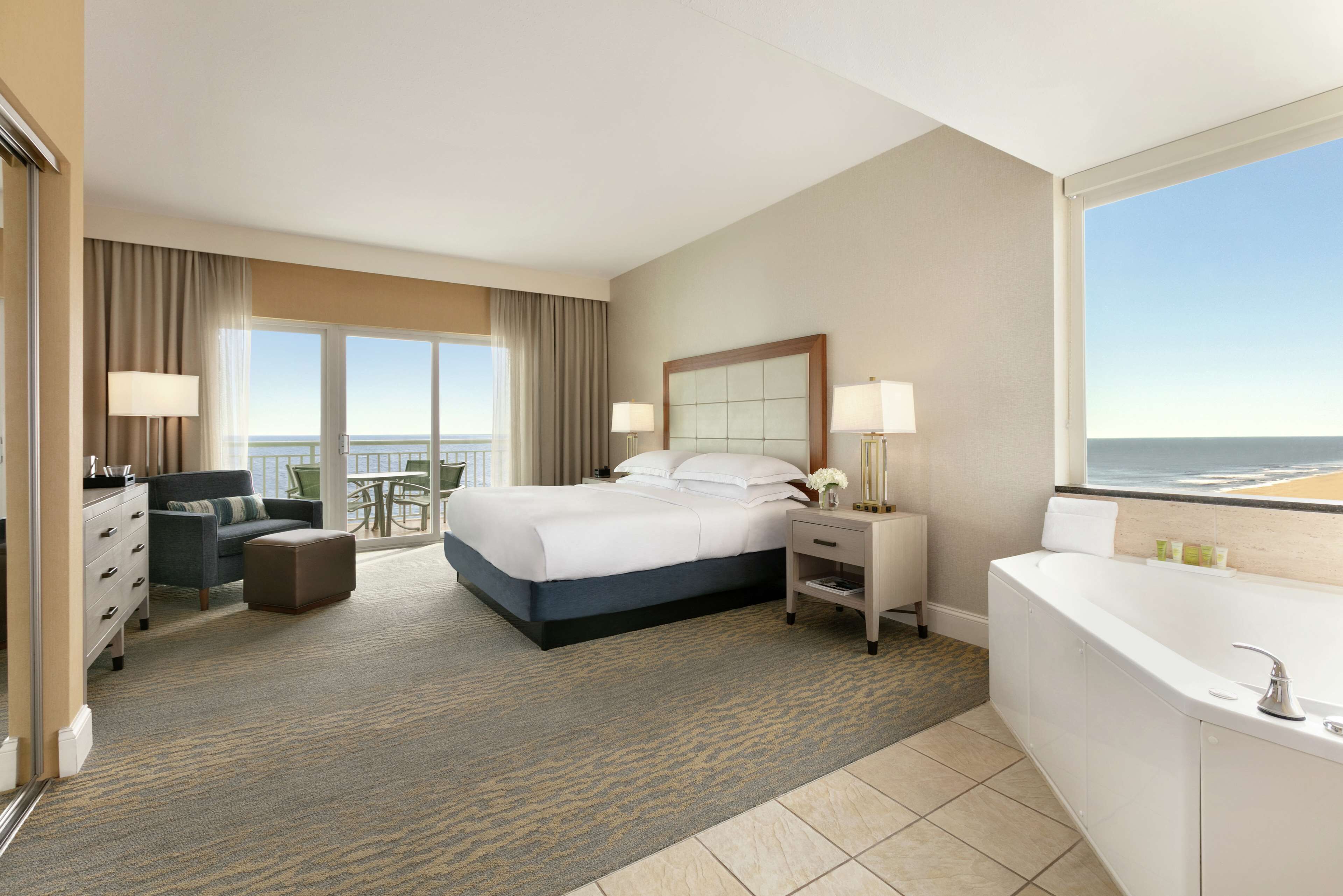A hotel room with a large bed, a sitting area with a chair and a small table, a bathroom with a bathtub, and a balcony with a view of the ocean.