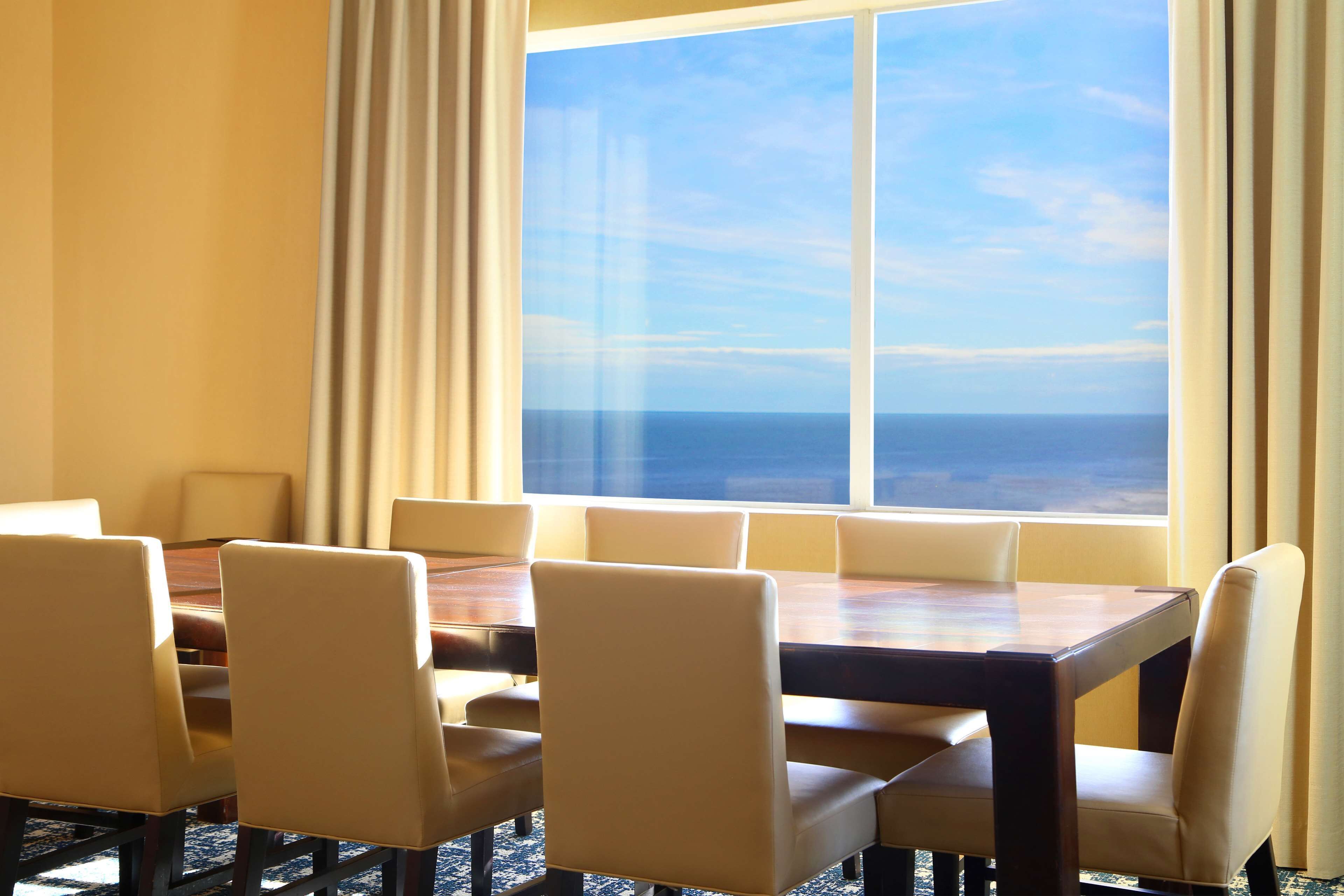 A conference room with a large table and six chairs, a large window with a view of the ocean, and beige curtains.