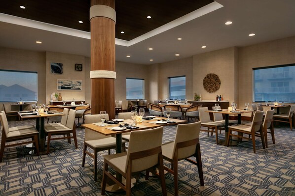 A hotel dining area with multiple tables and chairs, large windows, and a patterned carpet.