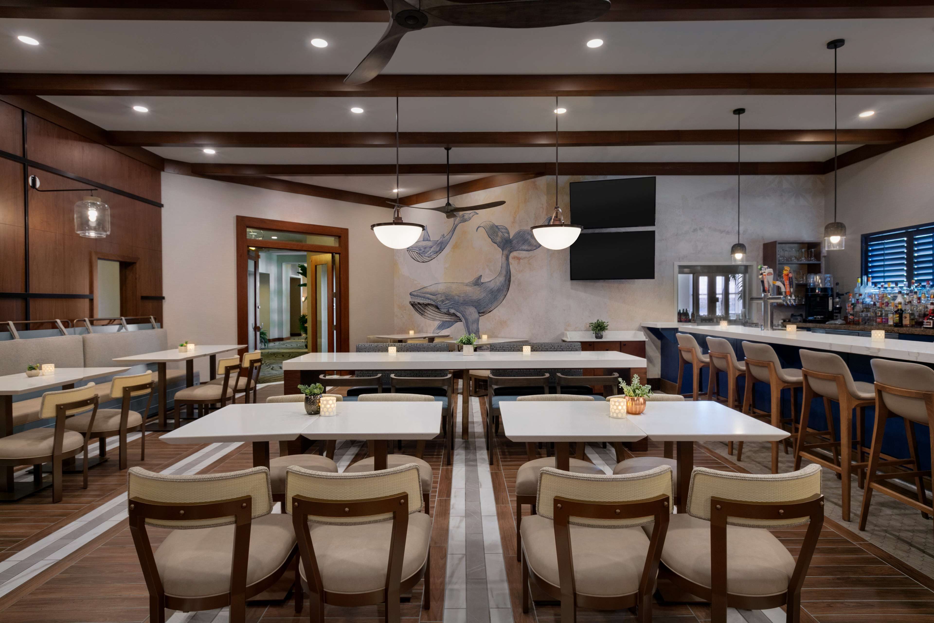 A restaurant with multiple tables and chairs, a mural of a whale, ceiling fans, and a bar area.