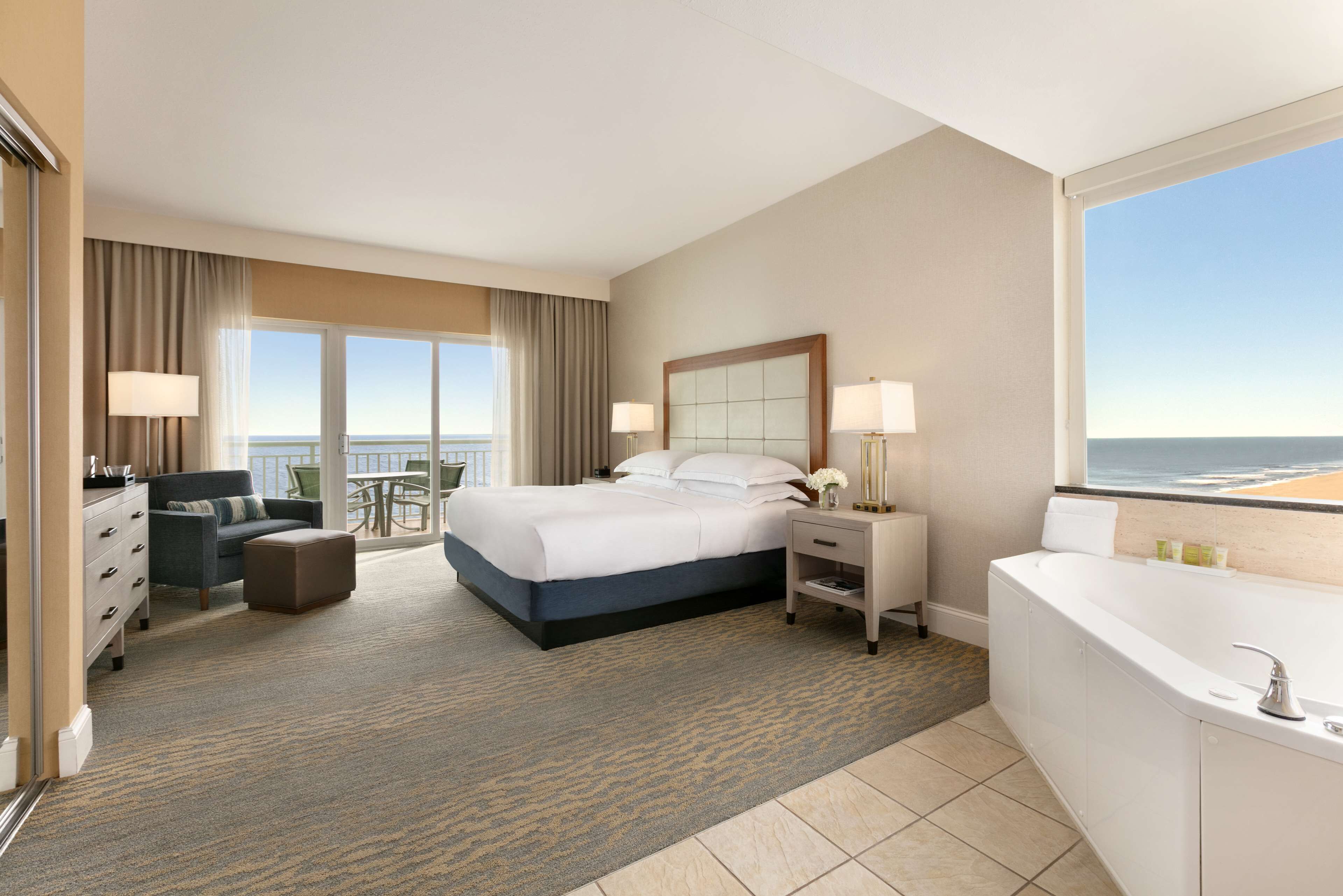 Habitación de hotel con una cama grande, un área de descanso con una silla y una mesita, un baño independiente con bañera y vistas a la playa desde la ventana.